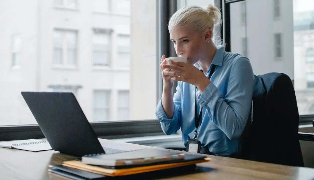 Mujer en camisa azul bebiendo una taza de café mientras trabaja en su computadora portátil en una oficina brillante.