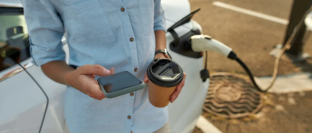 a woman charging her car while checking het phone and holding a take-away coffee cup