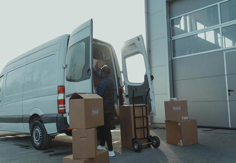 A man outside of a warehouse, placing some boxes in a white light-commercial vehicle