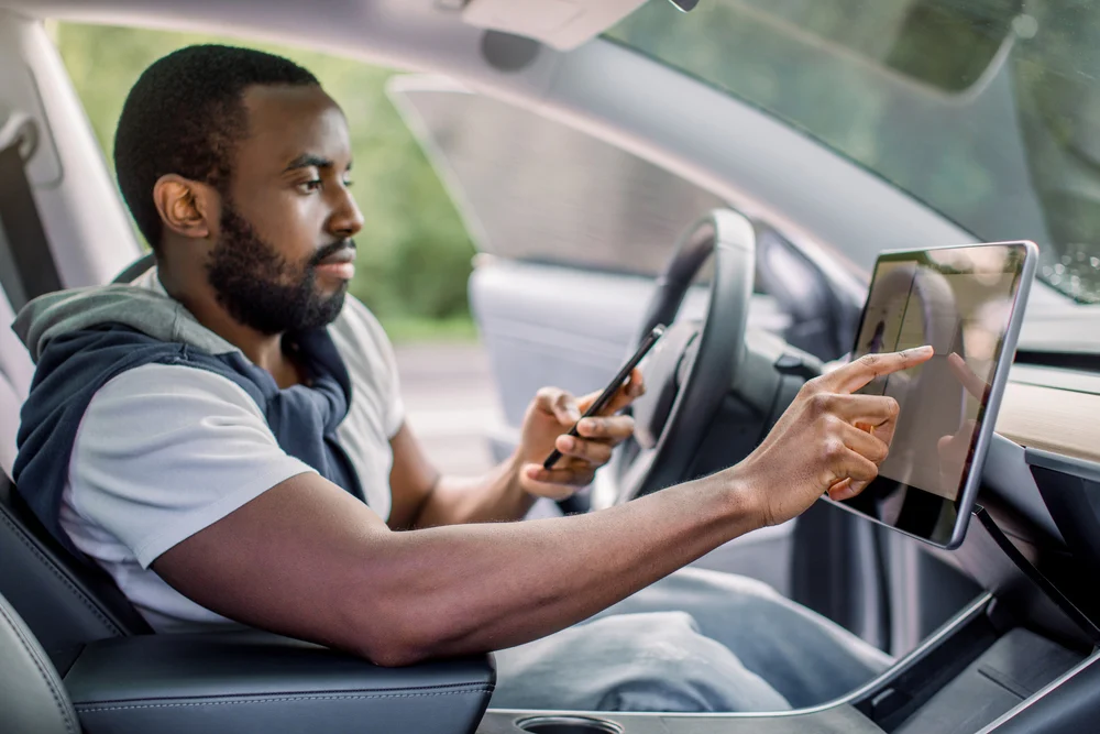 A man is sitting inside an electric car and is selecting some features on its digital dashboard, while holding his smartphone in the other hand.