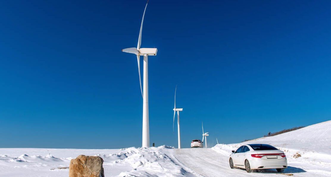 En tesla som kjører på en snødekt vei med blå himmel og vindturbiner