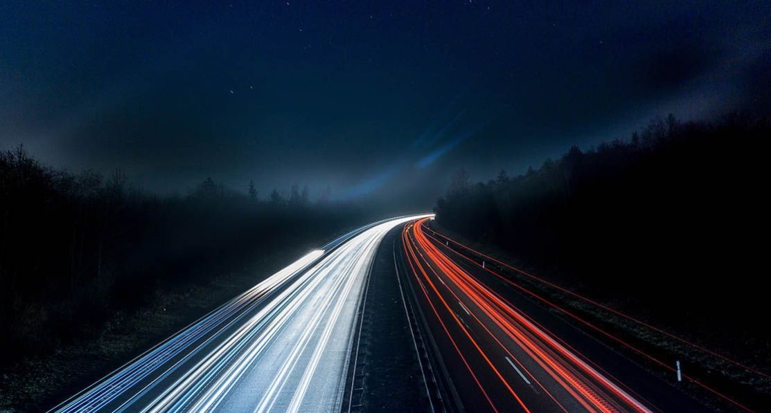 A motorway at night in the middle of the woods with light beams that represent vehicles' speed.
