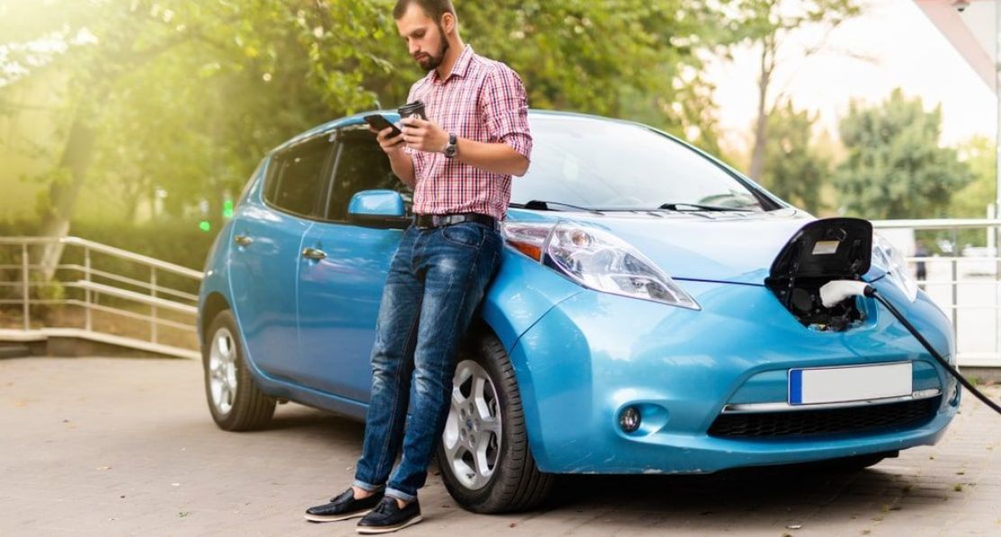 A man is checking his smart phone while leaning on his electric car whilst it is charging.