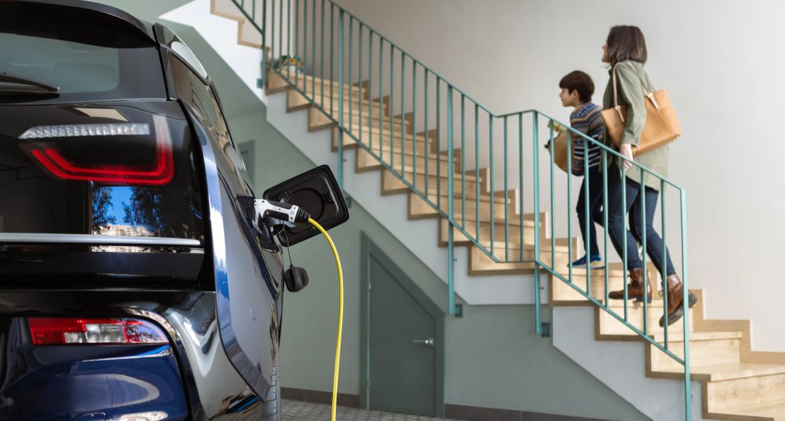A blue electric car charging in a garage. In the background, a woman and a child are climbing the stairs to enter the house.