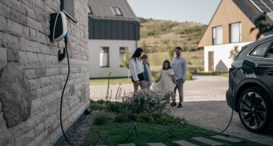 Ein elektrisches Auto ist mit einer EVBox Elvi Ladestation verbunden, die an eine Hauswand montiert ist. Eine Familie steht im Hintergrund und hat Spaß.
