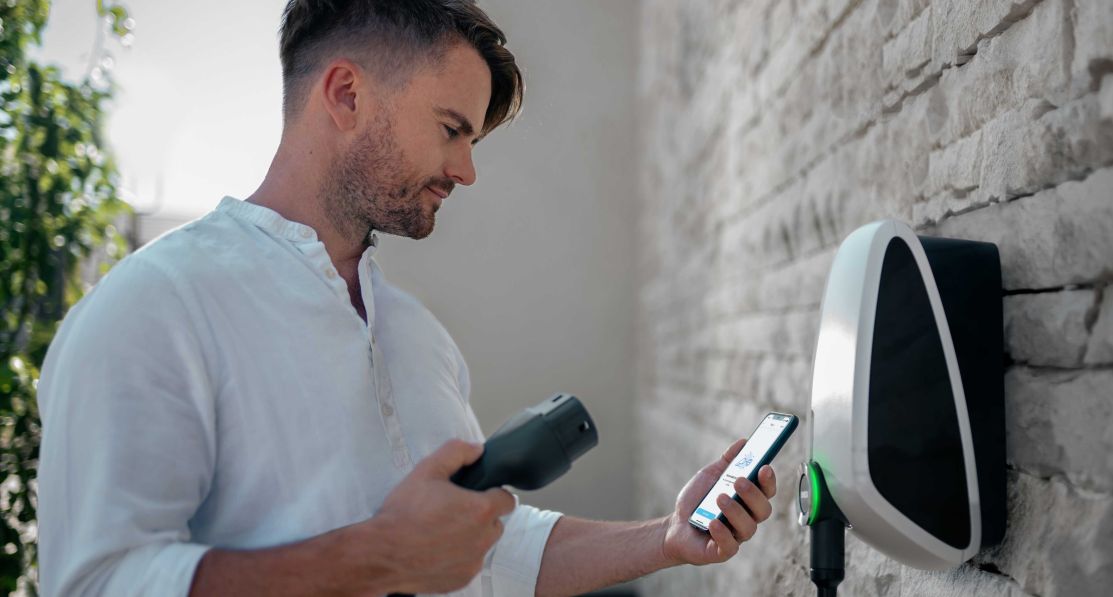 Un homme regardant tenant le câble de charge d'une main et son smartphone de l'autre, attendant de se connecter à la wallbox Elvi home.
