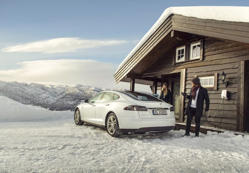 A woman and a man standing in front of a cabin in a wintery landscape looking at their white Tesla parked in front of the door. The man holds a charging cable of an EVBox Elvi home EV charger that's mounted to the wall.