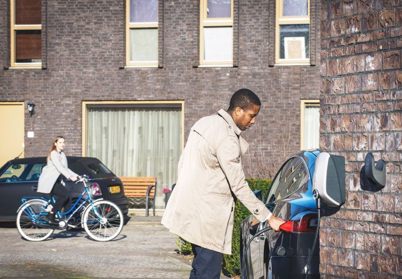 A homeowner plugging his Level 2 EVBox Elvi charger into his car, whilst a woman passes by on a bike and curiously looks at him.