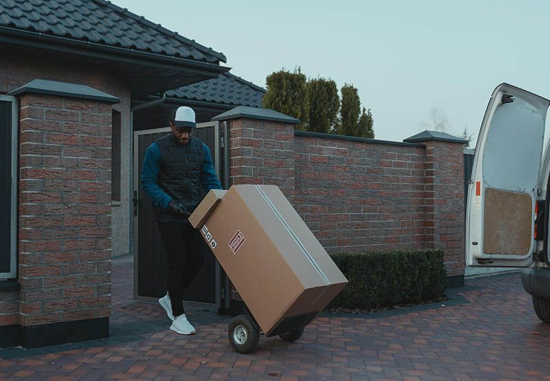Een bezorger duwt een karretje met een grote doos buiten een huis.