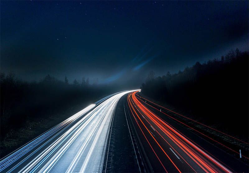A highway at night in the middle of the woods with light beams that represent vehicles' speed.