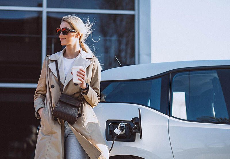 Een vrouw met een zonnebril op en casual gekleed, met een warm drankje in haar hand. Achter haar staat een auto opgeladen voor een modern gebouw.