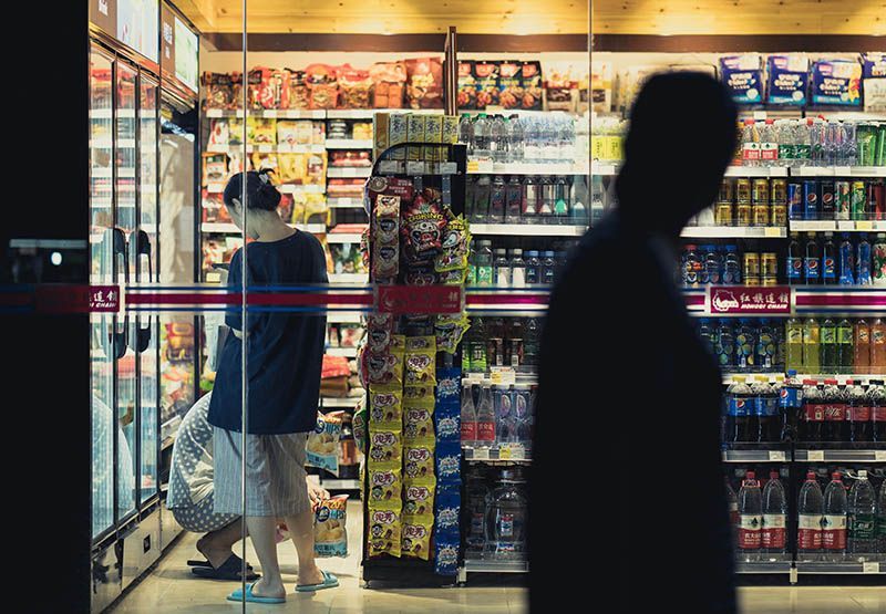 Two people in a convenience store at a fuel retail location are shopping groceries, while a person is walking outside the shop looking through the window.