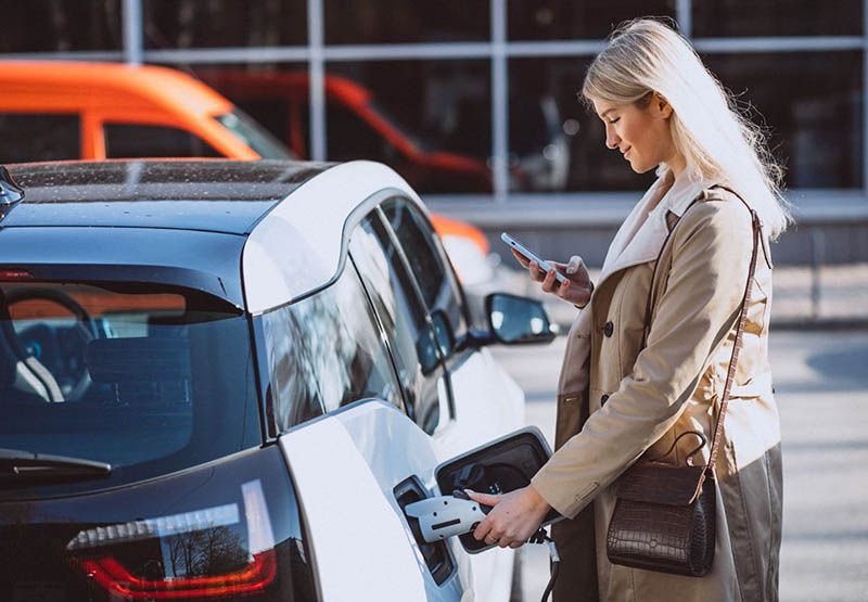 En kvinna i ledig klädsel tittar på sin smartphone medan hon laddar sin bil och lägger handen på laddningspluggen.