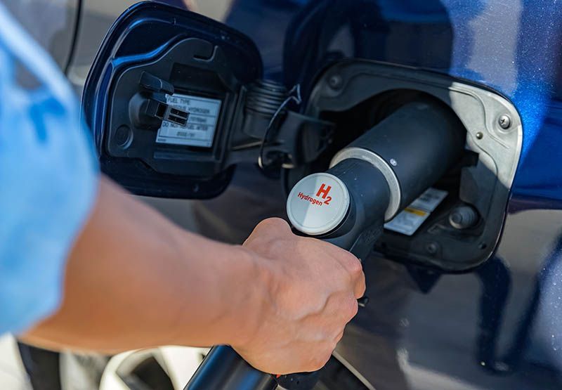 A person holding an Hydrogen pump to refuel a blue car.