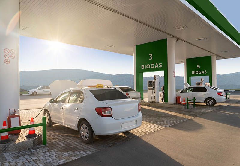 Two modern white cars are refuelling at a biogas station on a sunny day.