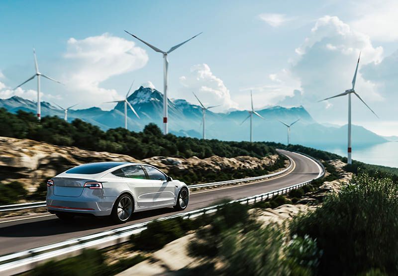 A modern white car is driving fast on a road with some wind turbines and mountains in the background on a sunny day.