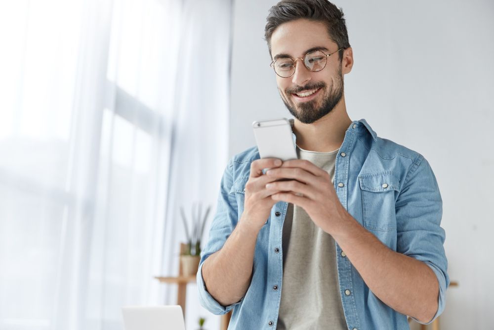 Ein bärtiger Mann mit Brille und gemütlicher Kleidung steht in seinem Wohnzimmer und lächelt sein Handy an, während er die wertvollen Infos liest, die er durch seine EV-Ladeapp bekommen hat.