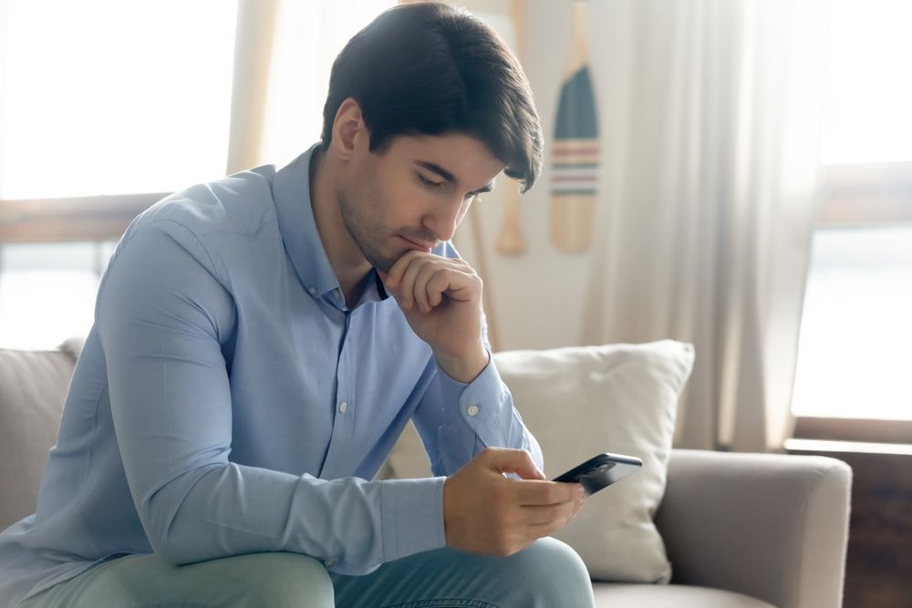 Ein Mann sitzt auf einem Sofa und schaut auf ein Handy, das er in seiner rechten Hand hält.