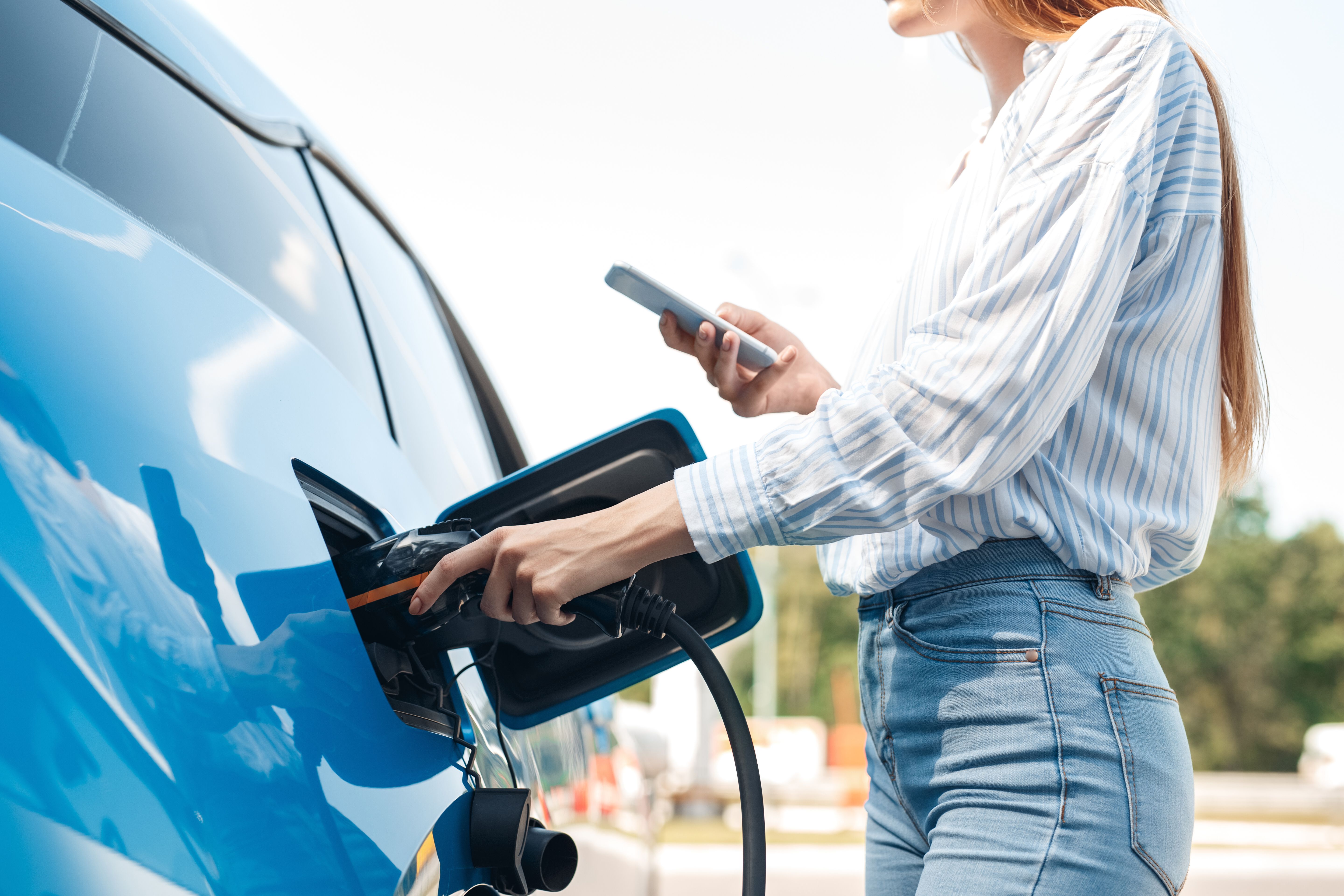 Eine Frau hält mit einer Hand ein Ladekabel, das an ihr Auto angeschlossen ist, während sie mit der anderen Hand ihr Telefon hält.