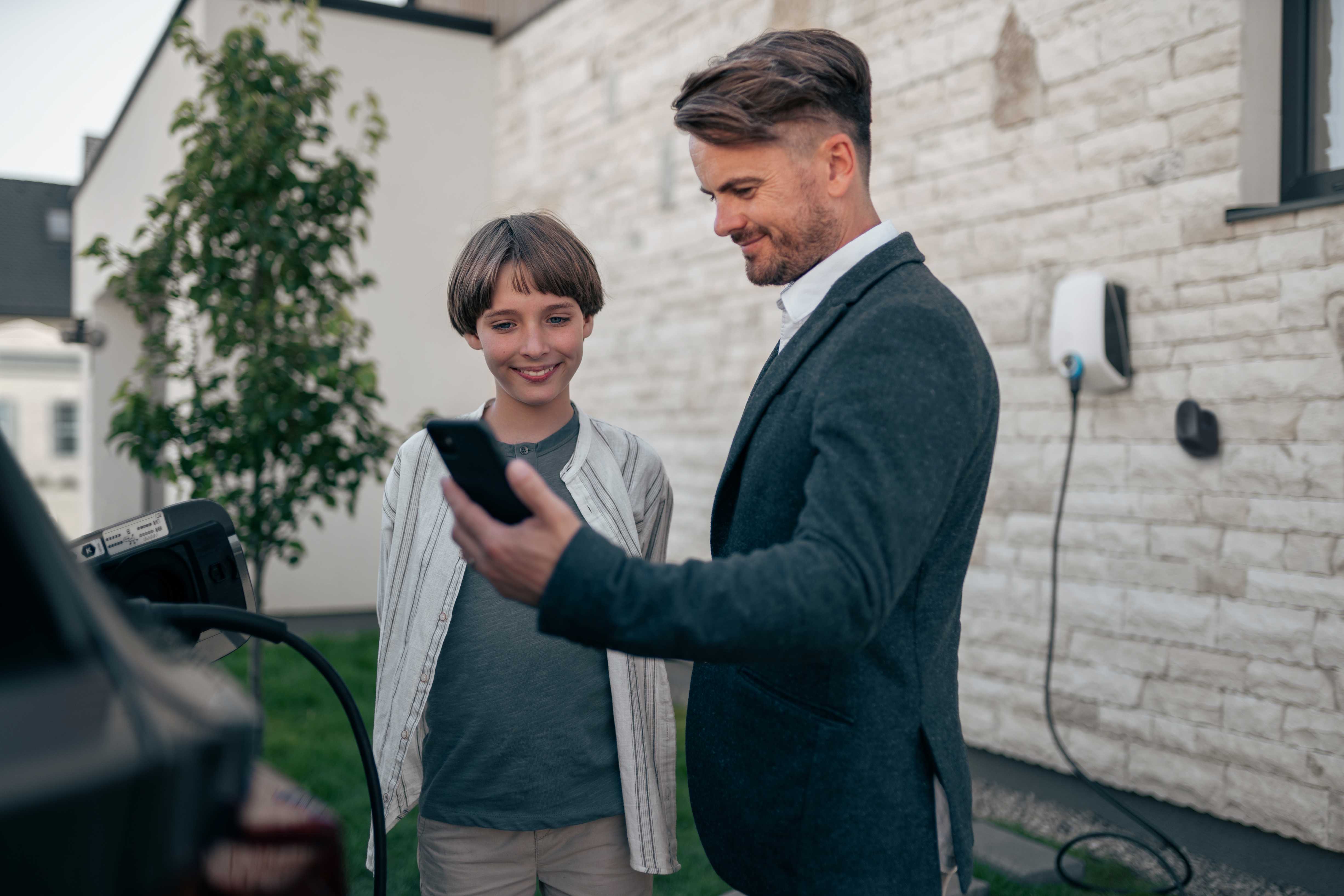 Ein Vater zeigt seinem Sohn sein Smartphone, das die App zum intelligenten Aufladen öffnet, während das Elektroauto aufgeladen und mit der an der Wand montierten Ladestation verbunden wird.