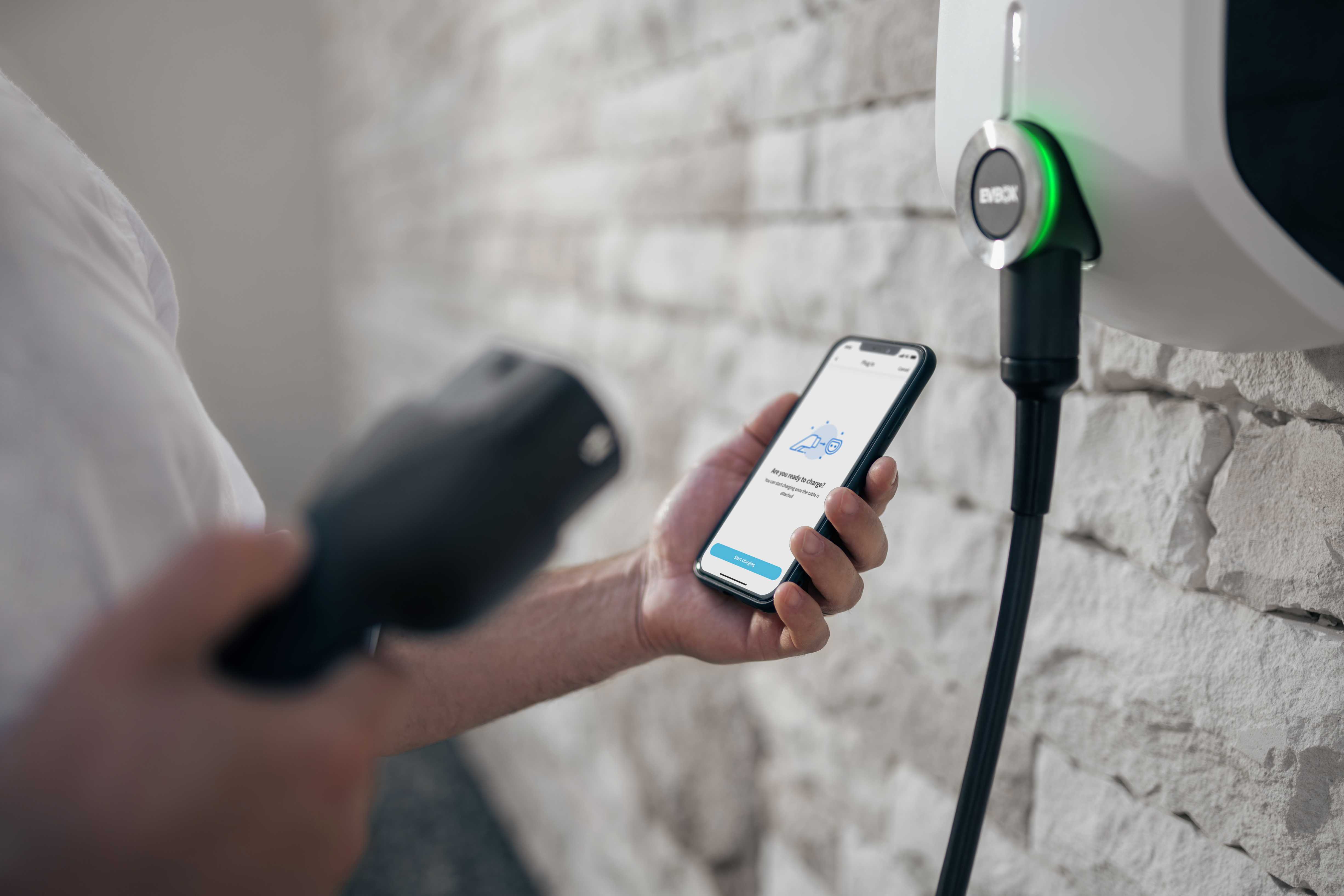 A man standing in front of an EVBox Elvi home charging station that's mounted on the wall. In one hand he's holding a smartphone on his left hand (showing the EVBox Everon charging app) with a message asking if he's are ready to charge. In his other hand, he holds the charging cable.