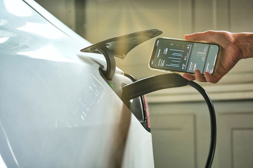 An EV connected to a charging station and a hand showing a smartphone with an app that shows data insights of the EV charging session.