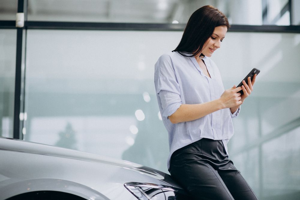 A woman sitting on the hood of her EV and looking at her phone while smiling.