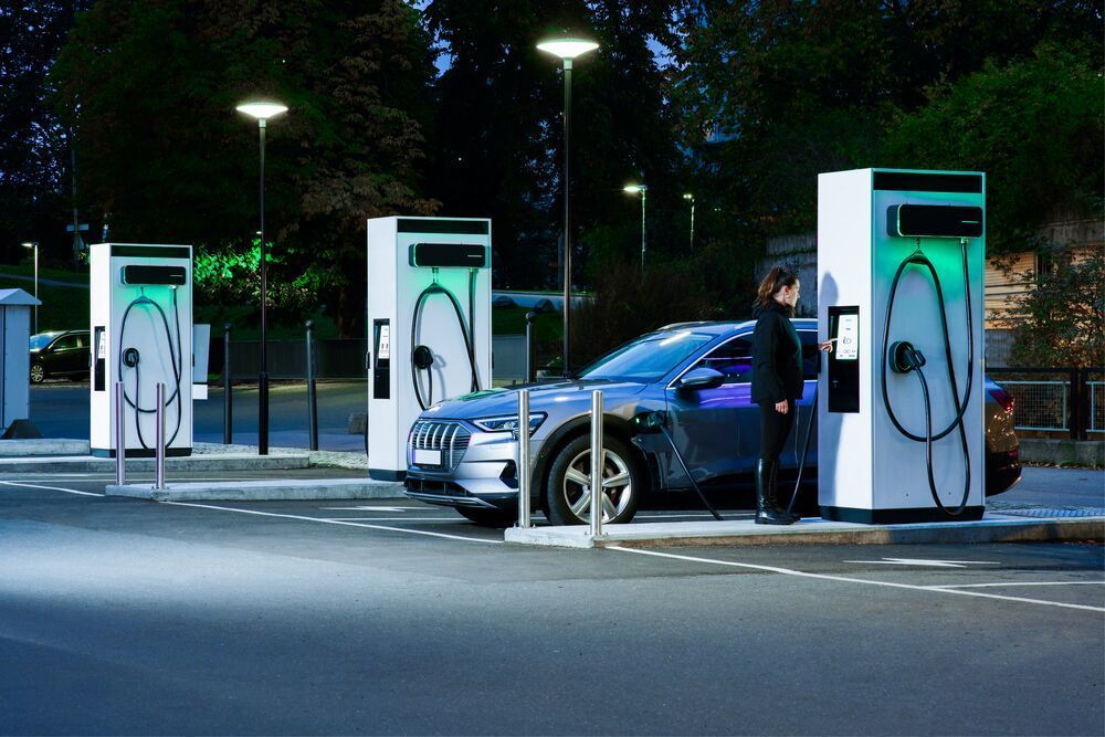 A parking lot with EVBox Troniq modular charging stations at night and a woman charging her EV on one. She's standing in front of the charging station and is checking out the screen of the charging station.