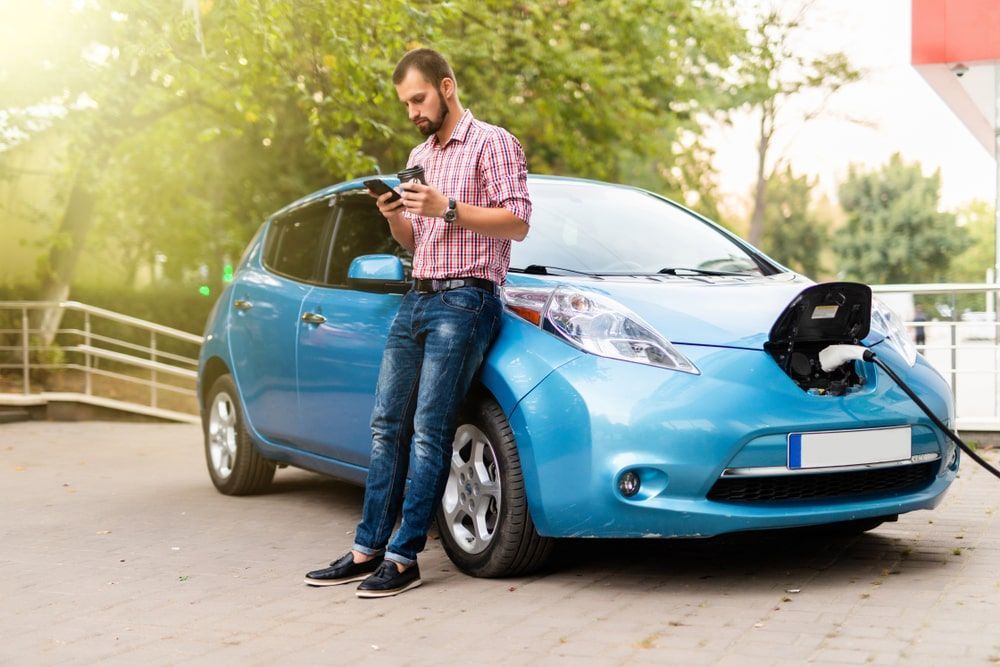 Man leaning against electric car getting charged looking at phone while holding a coffee.