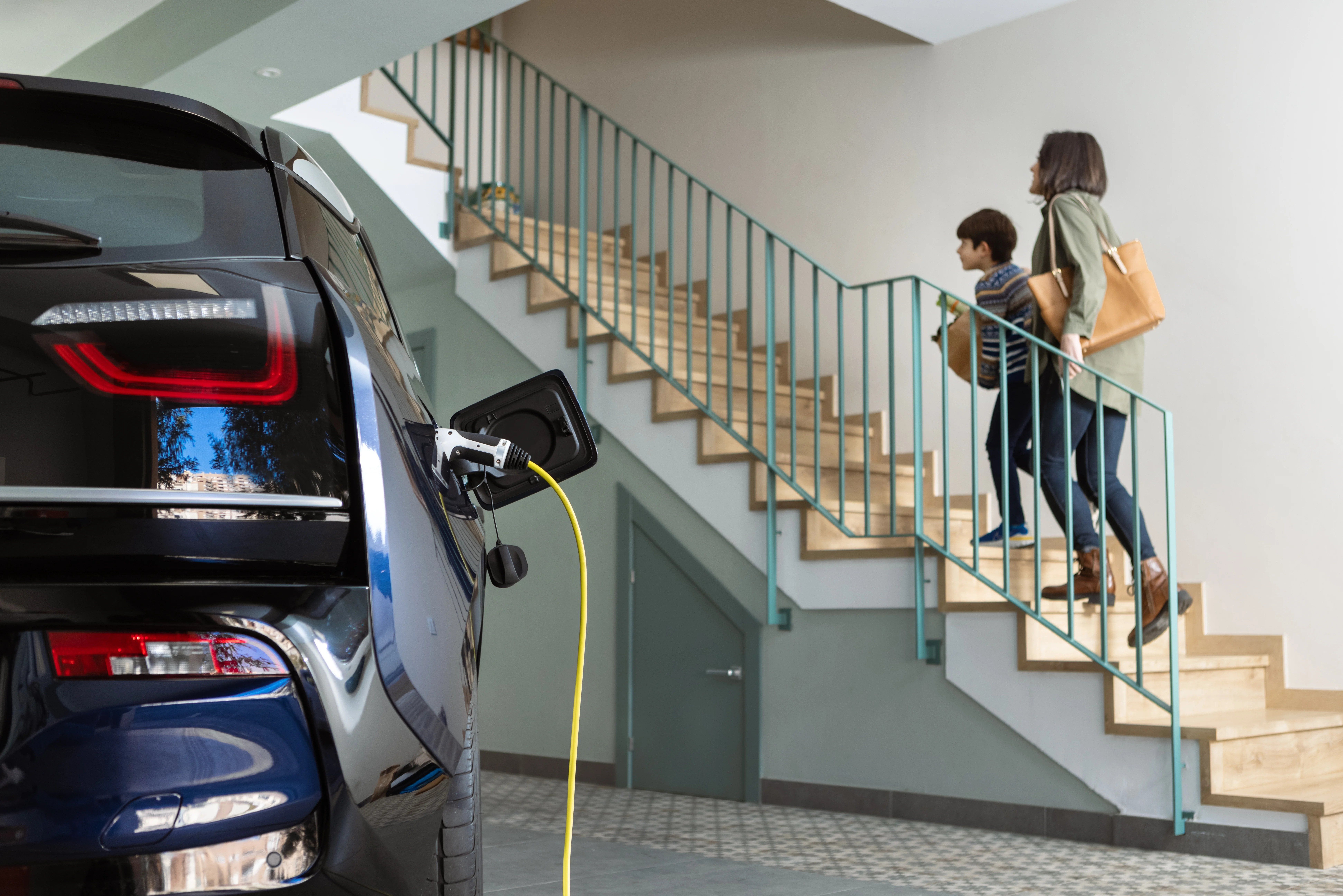 A blue electric car charging in a garage. In the background, a woman and a child are climbing the stairs to enter the house.