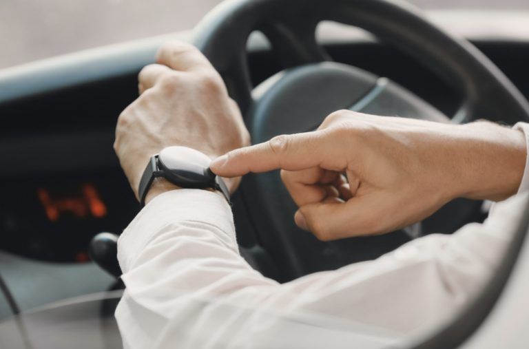 A zoomed in shot of 2 hands of a man behind the wheel of a parked car who's pointing at his watch.
