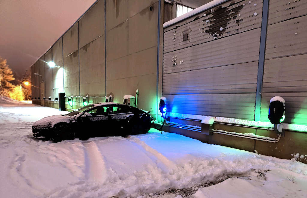 En Tesla på en parkeringsplass fylt med snø som lades ved en av mange EVBox BusinessLine AC-ladestasjoner montert på veggen om natten.