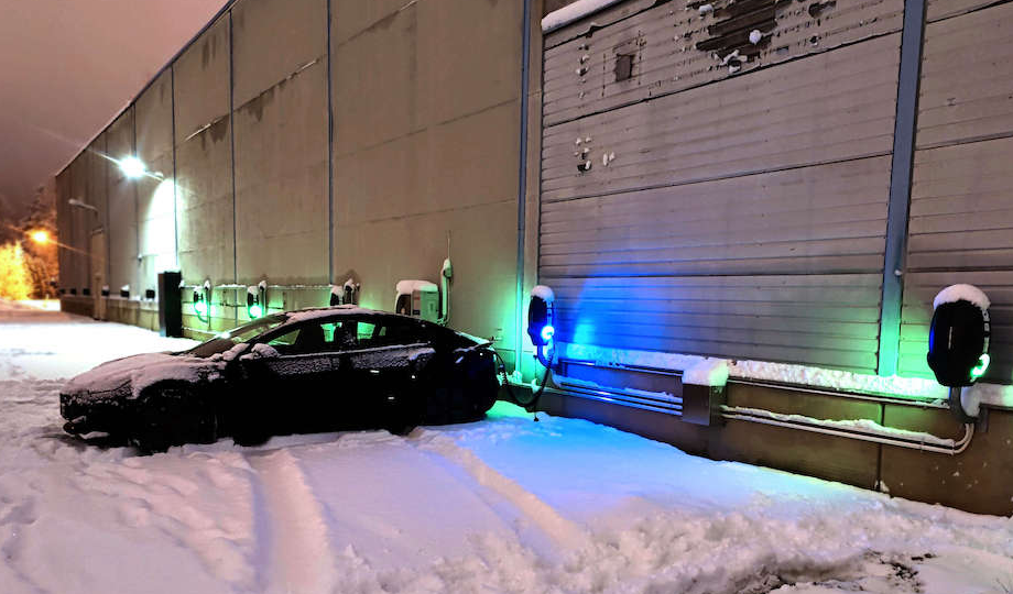 En Tesla på en parkeringsplass fylt med snø som lades ved en av mange EVBox BusinessLine AC-ladestasjoner montert på veggen om natten.