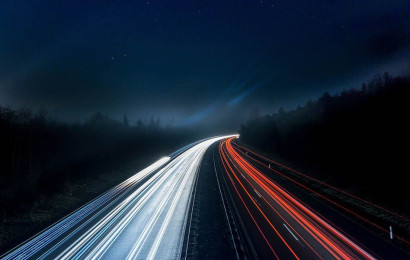 A highway at night in the middle of the woods with light beams that represent vehicles' speed.