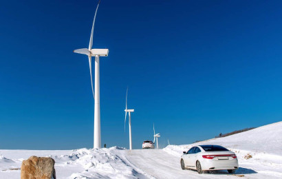 En Tesla som kjører på en snødekt vei med blå himmel og vindturbiner