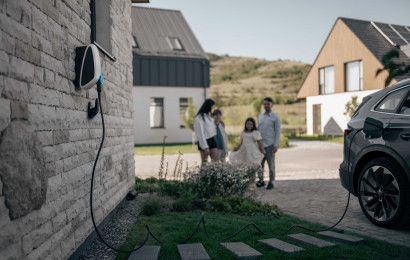An electric car is connected to the Elvi home charging station mounted at wall. A family is seen at the background having fun.