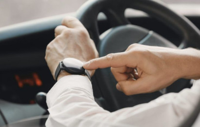 A zoomed in shot of 2 hands of a man behind the wheel of a parked car who's pointing at his watch.