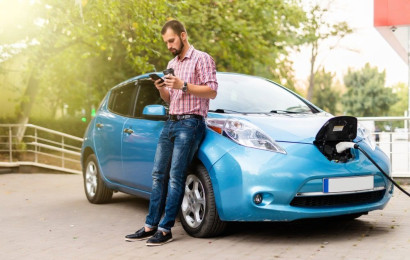 A man is checking his smart phone while leaning on his electric car whilst it is charging.